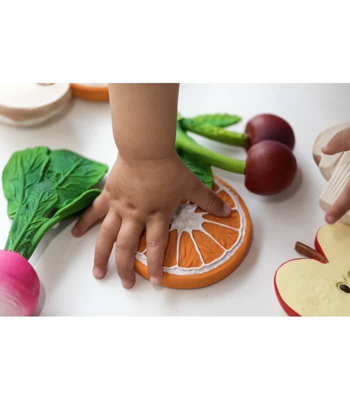 foto de la mano de un niño acercandose a clementino the orange mordedor para bebes de Oli y Carol rodeada de otras frutas de la coleccón fruits and veggies de la marca